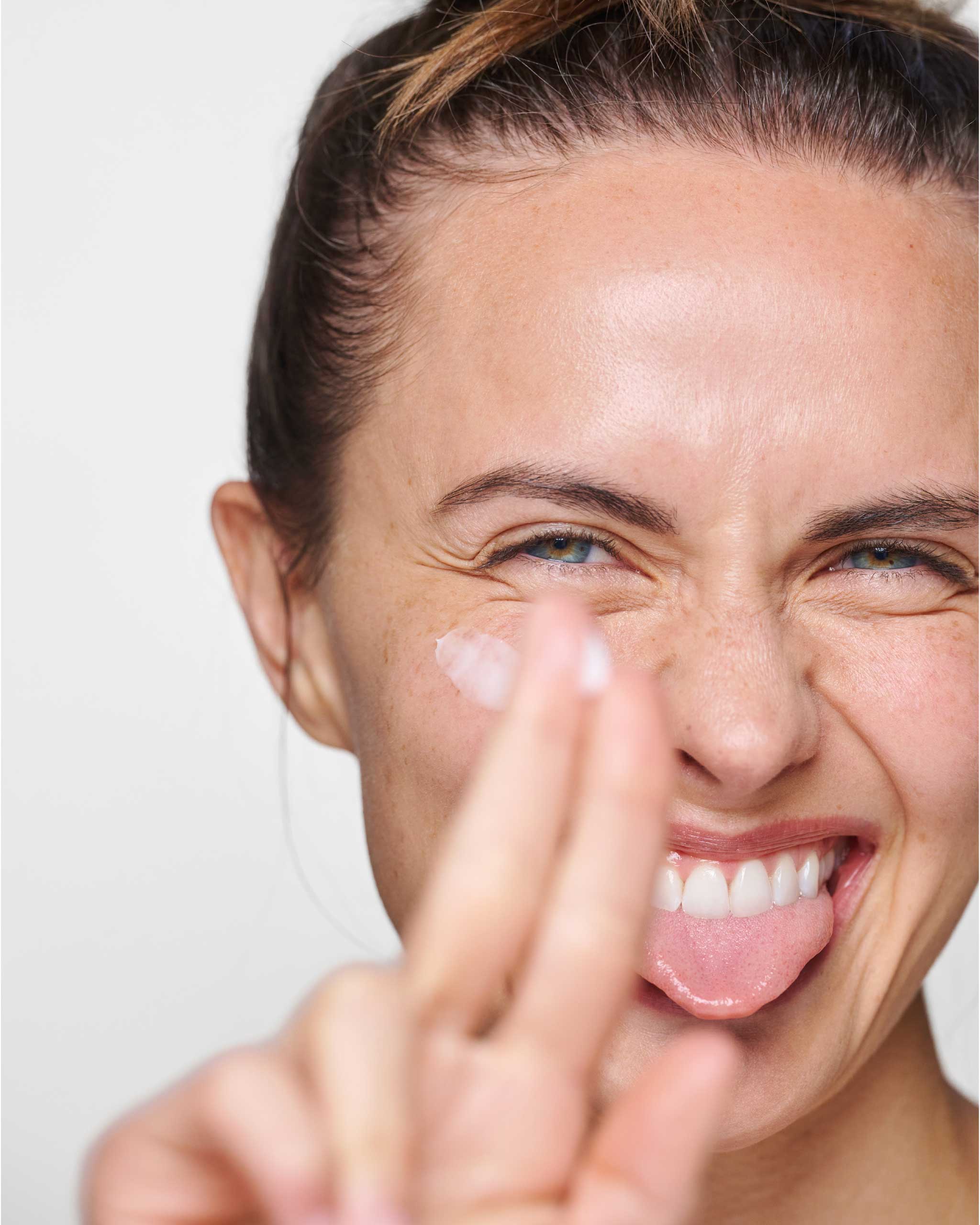 Smiling woman with tongue between teeth and Eye Cream on cheek & fingertips